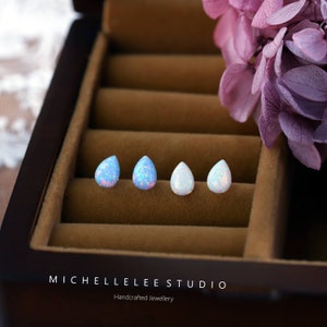 May include: Four pairs of teardrop-shaped opal earrings in a jewelry box. Two pairs are light blue with pink and white flecks, and two pairs are white with iridescent flecks. The box is brown with a white label that reads "MICHELLELEE STUDIO Handcrafted Jewellery".