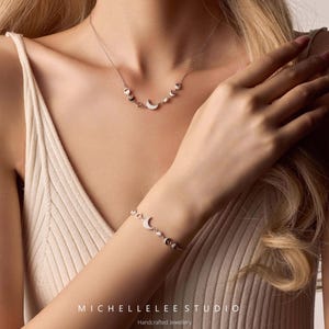 May include: A silver necklace and bracelet set featuring crescent moon and pearl accents. The jewelry is displayed on a person wearing a cream-colored tank top. The brand name "MICHELLELEE STUDIO" and the text "Handcrafted Jewellery" are visible.