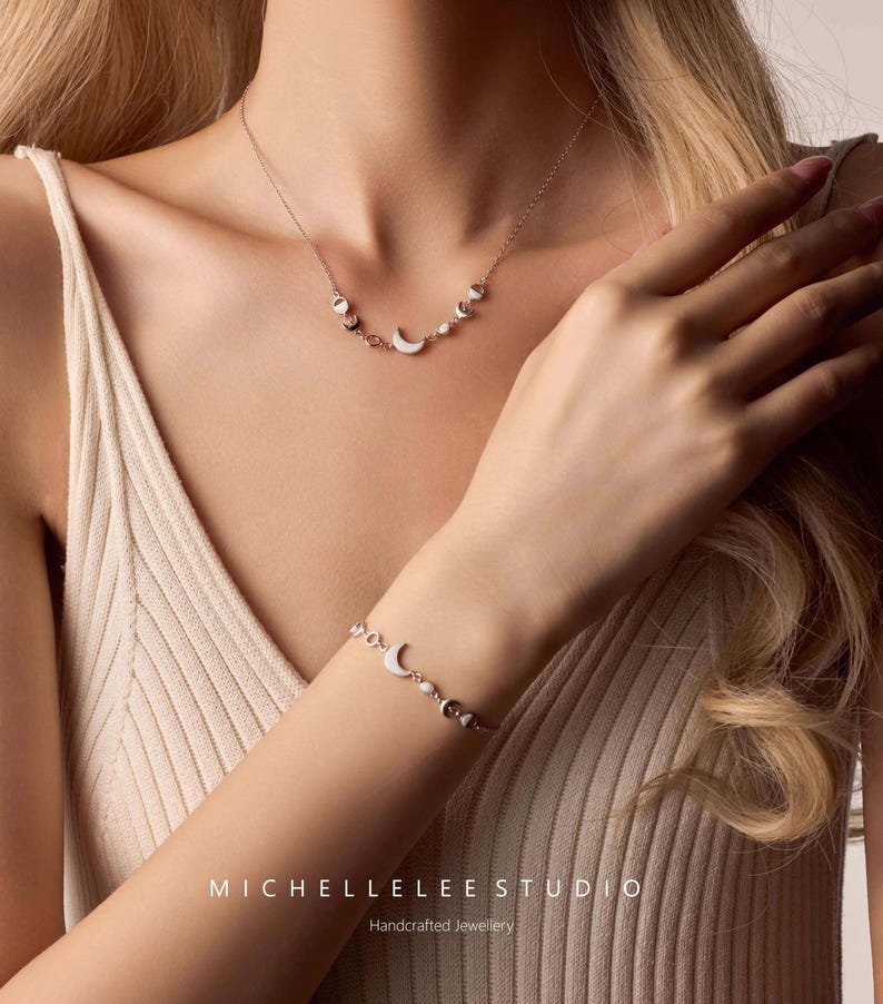 May include: A silver necklace and bracelet set featuring crescent moon and pearl accents. The jewelry is displayed on a person wearing a cream-colored tank top. The brand name "MICHELLELEE STUDIO" and the text "Handcrafted Jewellery" are visible.