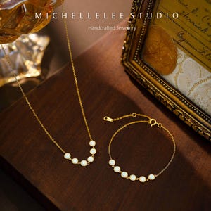 May include: A gold-toned necklace and bracelet set. The necklace and bracelet feature a series of white, oval-shaped stones. The image also includes the text "MICHELLELEE STUDIO" and "Handcrafted Jewellery". The jewelry is displayed on a dark brown surface.