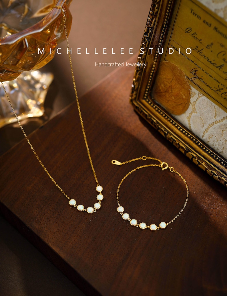 May include: A gold-toned necklace and bracelet set. The necklace and bracelet feature a series of white, oval-shaped stones. The image also includes the text "MICHELLELEE STUDIO" and "Handcrafted Jewellery". The jewelry is displayed on a dark brown surface.