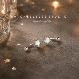 Könnte beinhalten: Zwei silberne, gebogene Barbell-Ohrringe mit sternförmigen Fassungen und irisierenden, weißen, opalartigen Steinen. Der Text MICHELLELEE STUDIO Handcrafted Jewellery ist oben sichtbar. Die Ohrringe haben kleine Kugeln an den Enden.