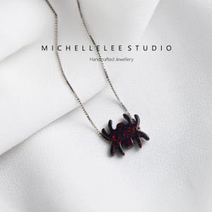May include: A silver chain necklace with a red glitter spider pendant. The pendant is a simple silhouette of a spider with eight legs. The necklace is on a white background.  Michelle Lee Studio Handcrafted Jewellery.
