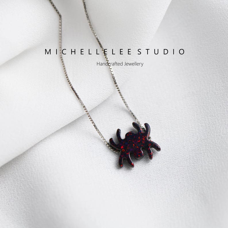 May include: A silver chain necklace with a red glitter spider pendant. The pendant is a simple silhouette of a spider with eight legs. The necklace is on a white background.  Michelle Lee Studio Handcrafted Jewellery.