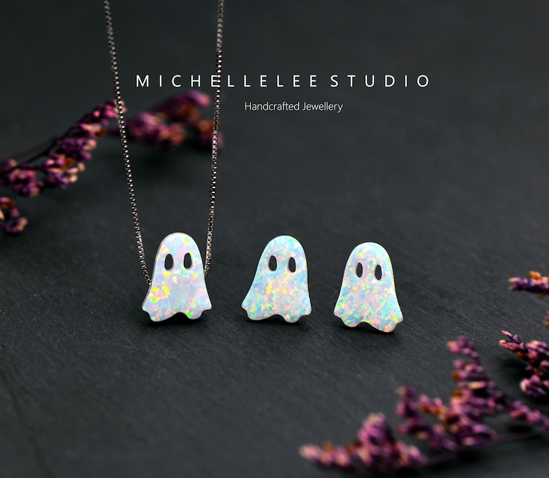 May include: Two silver necklaces with opal ghost charms. The ghosts are white with iridescent opal accents. The necklaces are on a black background.
