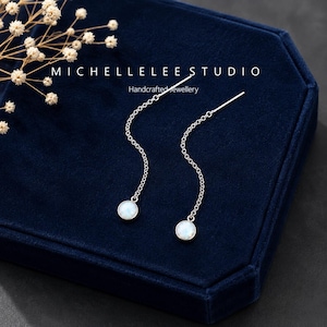 May include: A pair of silver threader earrings featuring a delicate chain and a small, round, iridescent white stone at the end. The text MICHELLELEE STUDIO Handcrafted Jewellery is visible above the earrings, which are displayed on a dark blue surface.