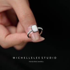 Puede incluir: Un anillo de plata con una piedra de ópalo blanco rectangular. La banda está adornada con pequeñas piedras preciosas transparentes. El anillo se muestra sobre un fondo negro. El texto en la parte inferior dice "MICHELLELEE STUDIO" y "Handcrafted Jewellery."