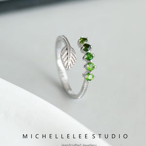 Può includere: Anello d'argento con un design a foglia e cinque pietre preziose verdi. L'anello è aperto e regolabile.