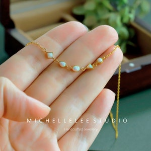 May include: A delicate gold chain necklace with a geometric design featuring a small square, oval, and triangle shapes accented with white opal stones. The necklace is displayed on a hand against a wooden background.