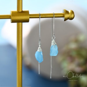 May include: A pair of silver chain earrings with light blue aquamarine crystal drops. The earrings are hanging from a gold stand. The text "mi Chellee Handmade Jewellery" is visible in the bottom right corner of the image.