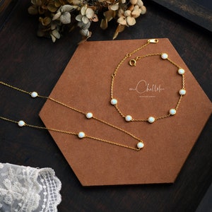 Pode incluir: Um conjunto de colar e pulseira dourados com pequenas pedras brancas redondas. As joias s&atilde;o exibidas em uma superf&iacute;cie hexagonal marrom, com flores secas ao fundo. A marca "mr. Chellelee" &eacute; vis&iacute;vel.