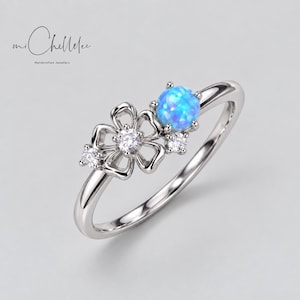 May include: A silver-toned ring featuring a central flower-shaped motif with open petals, a round bright blue opal, and three small clear round stones. The band is smooth and polished, and the text mi Chellelee Handcrafted Jewellery is visible.