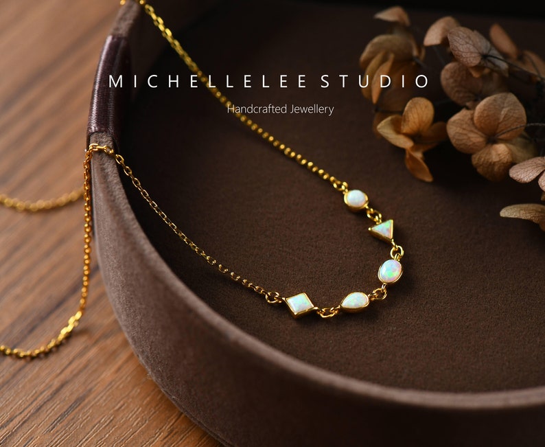 May include: A gold chain necklace with white opal stones in various shapes, including ovals, squares, and triangles. The necklace is displayed in a brown velvet box with the text "MICHELLE LEE STUDIO Handcrafted Jewellery".
