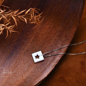 May include: A silver chain necklace with a white ceramic pendant featuring a star cutout. The pendant is on a brown wooden surface.