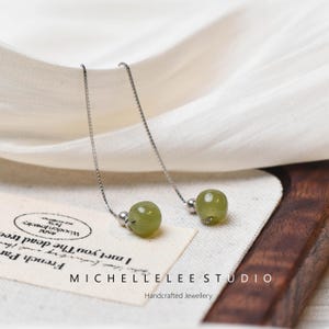 Op de afbeelding: Een paar zilveren oorhangers met groene jade stenen kralen. De oorhangers liggen op een witte stoffen achtergrond met een klein label waarop staat "I met you the dead tree Wooden Joucky French Par".