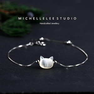 May include: A silver bracelet with a cat face pendant. The pendant is a light, pearly white colour, with two small silver beads either side. The bracelet is on a black surface. Text on the image reads: MICHELLELEE STUDIO Handcrafted Jewellery.