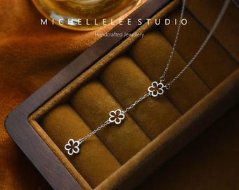 Puede incluir: Un delicado collar de plata con tres colgantes en forma de flor que cuelgan de una cadena. El collar se muestra en una caja de madera con forro de terciopelo.