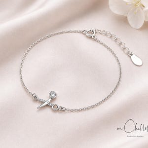 May include: A delicate silver bracelet featuring a scissors charm adorned with small clear crystals and a larger crystal. The bracelet has an adjustable chain and a lobster clasp. The background is a soft, light-colored fabric.