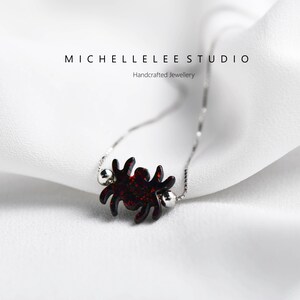 May include: A silver chain necklace with a red glitter spider pendant. The pendant is attached to the chain with two small silver beads. The necklace is on a white fabric background. MICHELLELEE STUDIO Handcrafted Jewellery.