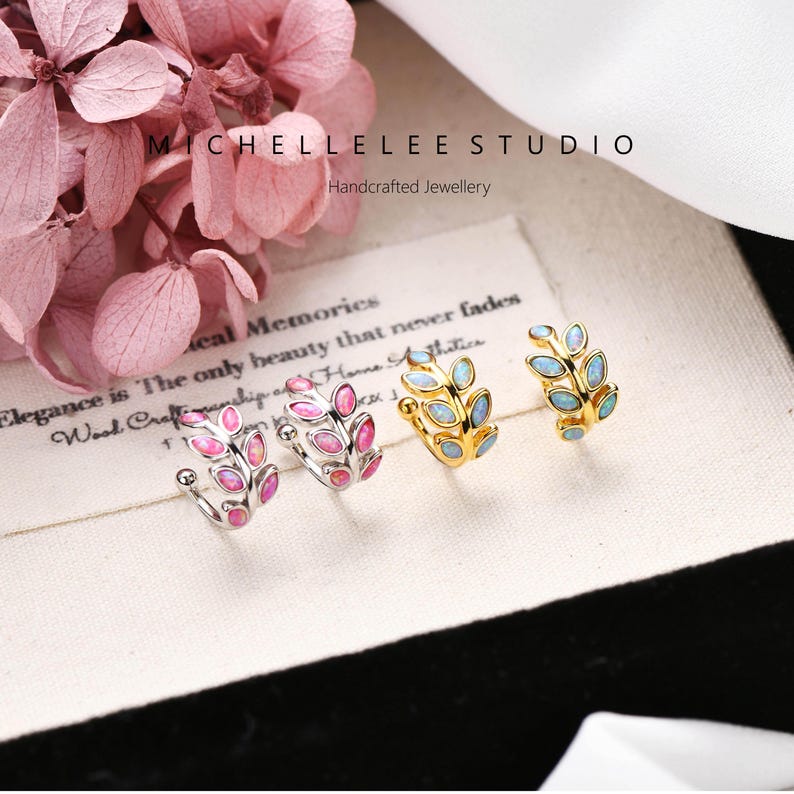 Puede incluir: Cuatro pendientes tipo ear cuff con forma de ramas de hojas, dispuestos en fila. Dos son de tono plateado con piedras ovaladas rosas y dos son de tono dorado con piedras ovaladas azul claro. El texto MICHELLELEE STUDIO Handcrafted Jewellery es visible.