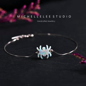 May include: A silver chain bracelet with a white opal spider charm. The bracelet is on a black background.