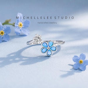 May include: A silver ring with a flower design. The ring features a large flower with blue opal petals and a smaller flower on the band. The image also includes the text "MICHELLELEE STUDIO Handcrafted Jewellery".
