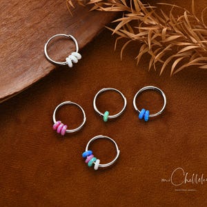 Può includere: Orecchini a cerchio in argento con accenti di perline colorate. Gli orecchini presentano piccoli cerchi rotondi con varie combinazioni di perline rosa, blu, verdi e bianche. Sono esposti su una superficie marrone con foglie secche.
