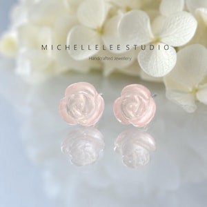 Può includere: Un paio di orecchini a lobo a forma di rosa rosa pallido con una finitura traslucida. Il testo MICHELLELEE STUDIO Handcrafted Jewellery è visibile sopra gli orecchini. Gli orecchini sono posizionati su una superficie riflettente con uno sfondo floreale bianco.