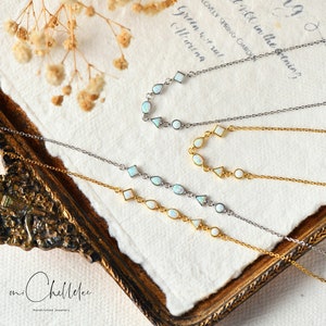 May include: Three delicate necklaces with opal stones and silver and gold chains. The necklaces are arranged on a white textured surface with the text "an Chellelee Handcrafted Jewellery".