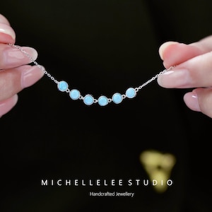 May include: A delicate silver necklace featuring seven light blue, round gemstones. The necklace is held against a dark background, showcasing the handcrafted jewelry. The text "MICHELLELEE STUDIO" and "Handcrafted Jewellery" are visible.