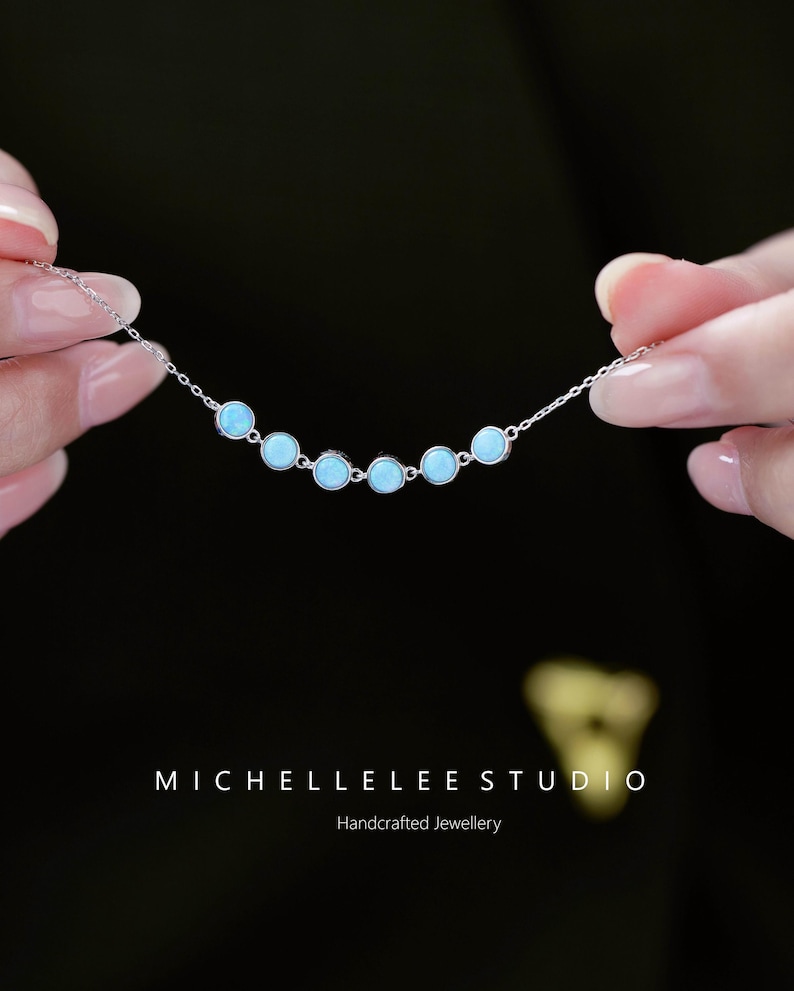 May include: A delicate silver necklace featuring seven light blue, round gemstones. The necklace is held against a dark background, showcasing the handcrafted jewelry. The text "MICHELLELEE STUDIO" and "Handcrafted Jewellery" are visible.