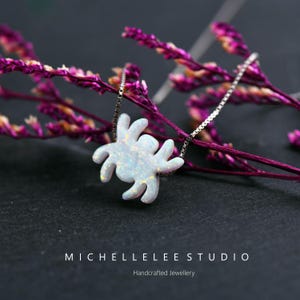 May include: A silver chain necklace with a white opal spider pendant. The pendant is a detailed, realistic depiction of a spider with eight legs. The necklace is on a pink floral background.