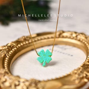 May include: A gold-colored chain necklace with a four-leaf clover pendant. The clover is a vibrant turquoise color. The necklace is displayed on a gold-framed surface. The text "MICHELLE LEE STUDIO Handcrafted Jewellery" is visible.