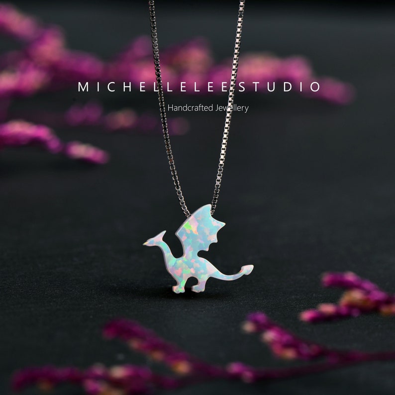 May include: A silver necklace with a dragon pendant. The pendant is white with iridescent accents, and the chain is a delicate silver. The background is dark, with the text "MICHELLELEEESTUDIO Handcrafted Jewellery".