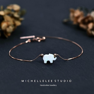May include: A delicate rose gold bracelet featuring a small, white opal elephant charm. The charm is centered on a fine chain, with small gold beads on either side. The bracelet is displayed against a dark background.