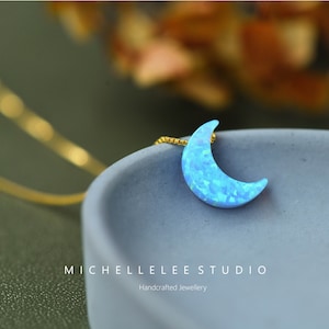 May include: A crescent moon pendant necklace with a gold chain. The moon pendant is a light blue opal with a textured surface. The necklace is displayed on a gray surface. The text "MICHELLELEE STUDIO" is visible.