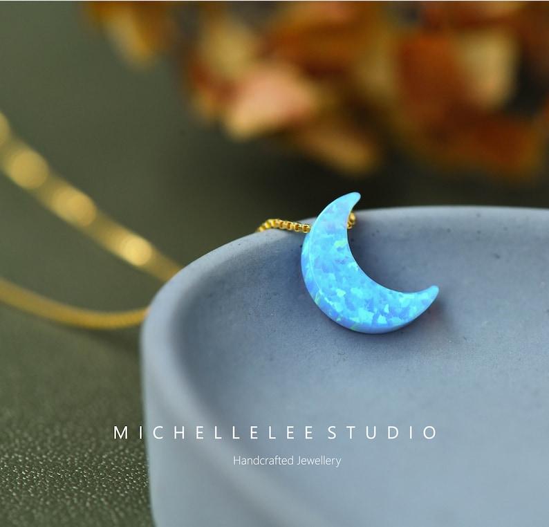 May include: A crescent moon pendant necklace with a gold chain. The moon pendant is a light blue opal with a textured surface. The necklace is displayed on a gray surface. The text "MICHELLELEE STUDIO" is visible.