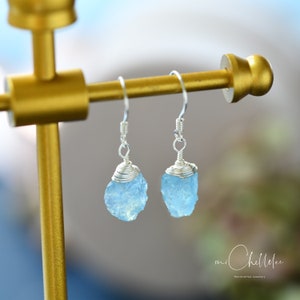 Puede incluir: Un par de pendientes de aguamarina envueltos en alambre plateado. Las piedras de aguamarina son de color azul claro y tienen una textura natural rugosa. Los pendientes están colgados de un soporte dorado.