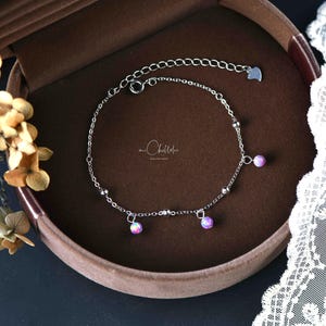 May include: A delicate silver bracelet with three small, round, iridescent pink beads. The bracelet is displayed in a brown jewelry box. The chain has a small heart-shaped charm at the end.