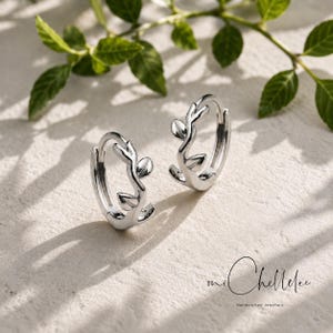 May include: A pair of silver hoop earrings featuring a delicate vine and leaf motif along the outer edge. The earrings have a polished metallic finish and a hinged closure mechanism. The text mi Chellelee Handcrafted Jewellery is visible.