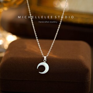 K&ouml;nnte beinhalten: Eine silberne Kettenkette mit einem Halbmond-Anh&auml;nger aus wei&szlig;em Perlmutt. Die Kette wird auf einer braunen Samtbox pr&auml;sentiert. Der Text "MICHELLELEE STUDIO Handgefertigter Schmuck" ist im Hintergrund sichtbar.