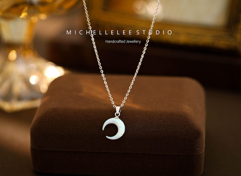 K&ouml;nnte beinhalten: Eine silberne Kettenkette mit einem Halbmond-Anh&auml;nger aus wei&szlig;em Perlmutt. Die Kette wird auf einer braunen Samtbox pr&auml;sentiert. Der Text "MICHELLELEE STUDIO Handgefertigter Schmuck" ist im Hintergrund sichtbar.