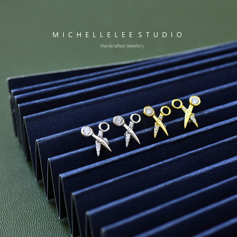 May include: Four pairs of silver and gold scissor earrings with a single clear rhinestone on each earring. The earrings are displayed on a blue background. MICHELLE LEE STUDIO Handcrafted Jewellery.