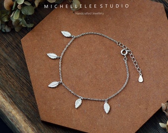 May include: A delicate silver bracelet featuring small leaf charms. The bracelet is displayed on a brown surface with the text "MICHELLELEE STUDIO Handcrafted Jewellery". The bracelet has an adjustable chain.