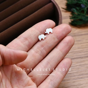 May include: A pair of small, white elephant stud earrings. The earrings are held in a hand, with the text "MICHELLELEE STUDIO Handcrafted Jewellery" visible below. The earrings have a textured surface and are set against a blurred background.