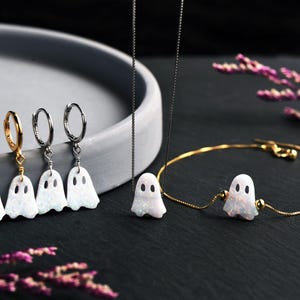 Pu&ograve; includere: Un set di gioielli composto da quattro orecchini a cerchio con ciondoli a forma di fantasma bianco iridescente, una collana a catena sottile con un ciondolo a forma di fantasma e un bracciale a catena dorata con un ciondolo centrale.