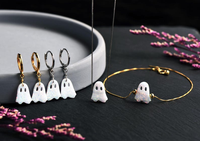 Pu&ograve; includere: Un set di gioielli composto da quattro orecchini a cerchio con ciondoli a forma di fantasma bianco iridescente, una collana a catena sottile con un ciondolo a forma di fantasma e un bracciale a catena dorata con un ciondolo centrale.
