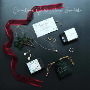 May include: An assortment of jewelry pieces, including necklaces, earrings, and rings, are arranged on a dark surface. A red ribbon and a sprig of greenery add decorative elements. The image includes the text "Christmas Gift Wrap Available".