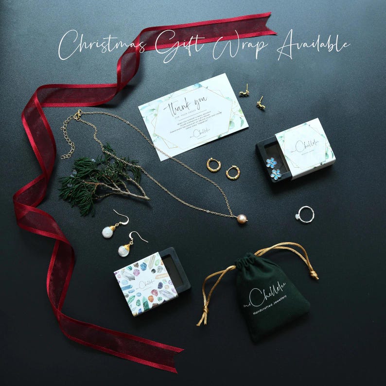 May include: An assortment of jewelry pieces, including necklaces, earrings, and rings, are arranged on a dark surface. A red ribbon and a sprig of greenery add decorative elements. The image includes the text "Christmas Gift Wrap Available".
