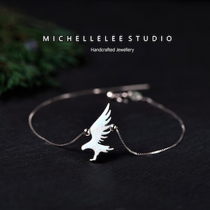 May include: A silver bracelet with a white eagle charm. The eagle has detailed wings and an iridescent finish. The bracelet is shown on a dark background. The image includes the text "MICHELLELEE STUDIO Handcrafted Jewellery".
